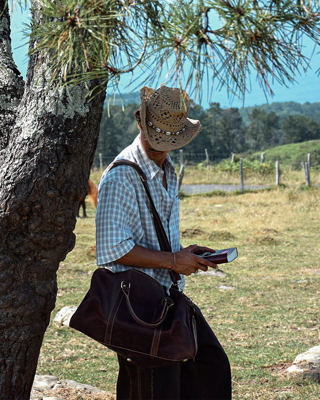 A man in a cowboy hat stands outdoors near a tree, wearing a blue and white plaid shirt and carrying a large dark brown leather duffel bag over his shoulder while holding a book.