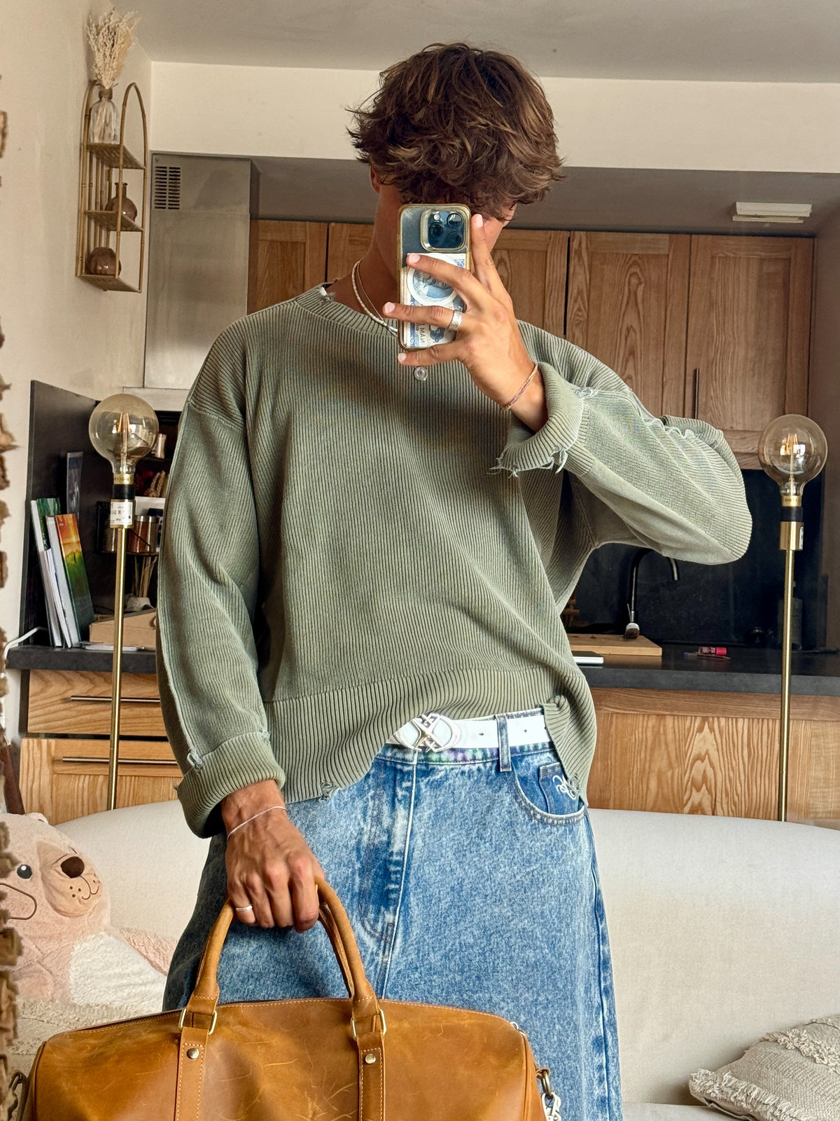 A person taking a mirror selfie in a cozy room, wearing an oversized sage green knit sweater and a blue denim skirt, holding a brown leather duffel bag.