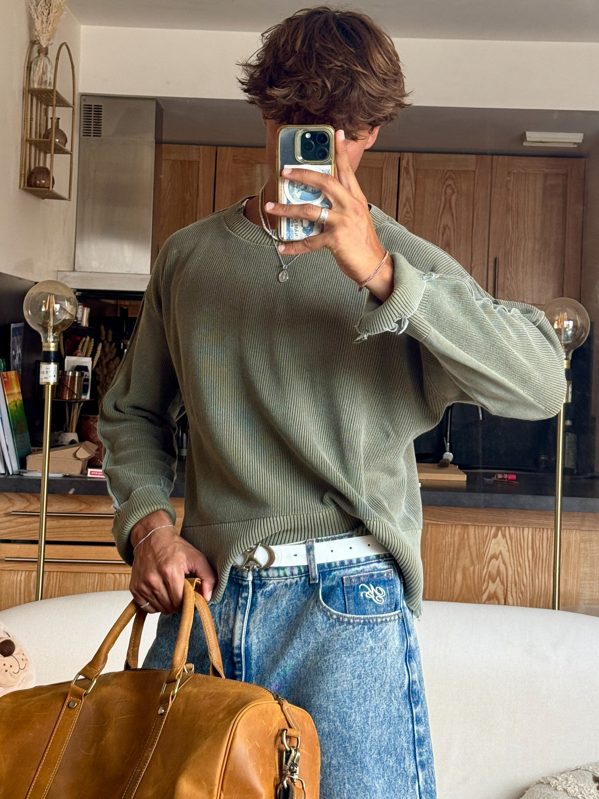 A person taking a mirror selfie in a living room setting, wearing a muted sage green knit sweater and blue jeans, holding a tan leather COFFT duffel bag.