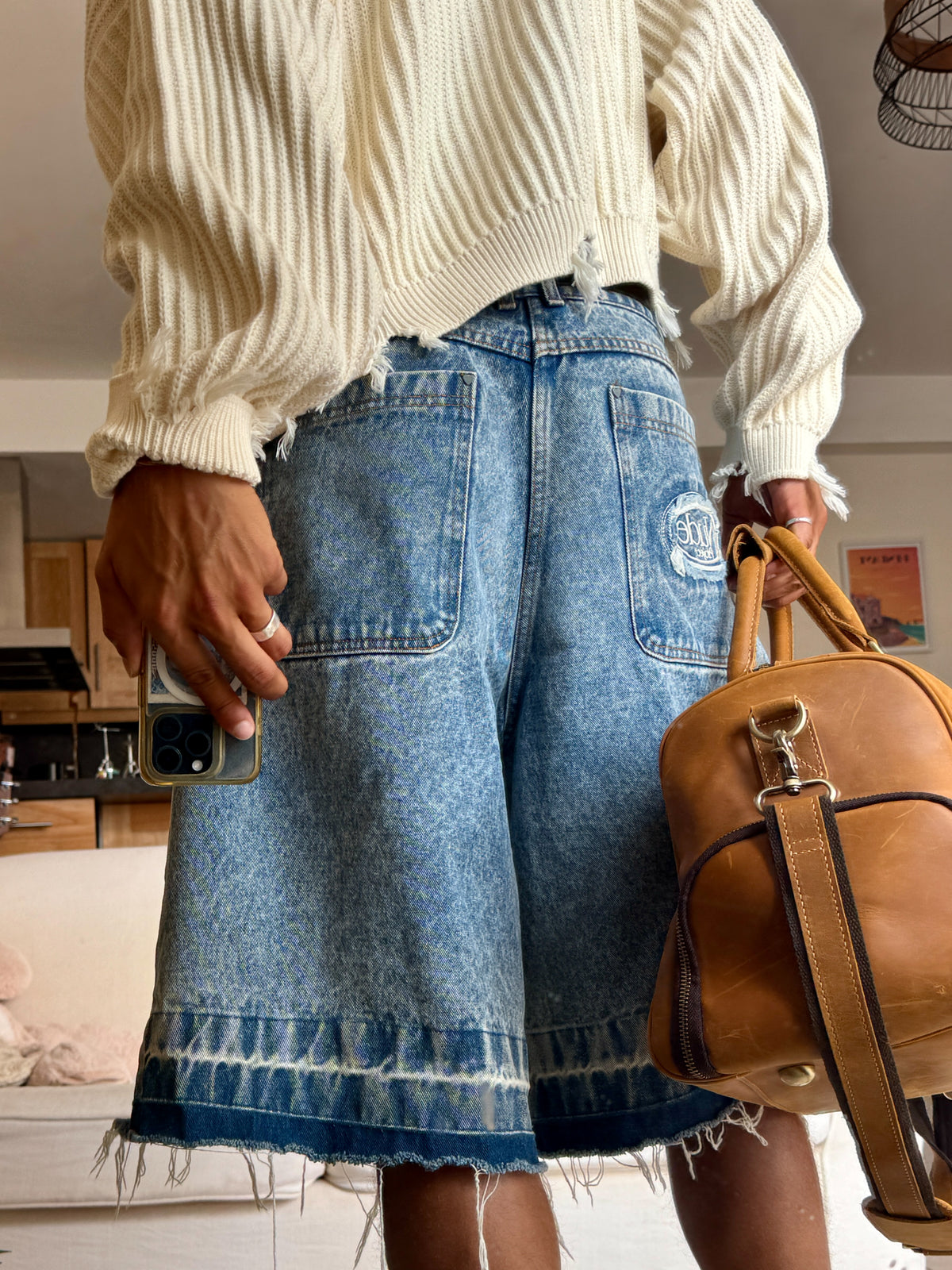 A person wearing light-wash blue denim shorts with a raw frayed hem and a cream ribbed knit sweater, holding a brown leather shoulder bag in an indoor setting.