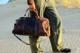 COFFT DUFFLE 01 Espresso leather duffle bag carried on sand dunes in warm outdoor lifestyle setting