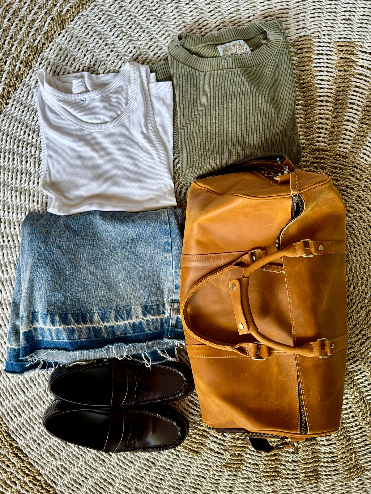 A styled flat-lay of folded clothing and a duffel bag on a woven straw surface. The set includes a white short-sleeve T-shirt, an olive green knit top, a light blue frayed-hem denim skirt, black leather loafers, and a tan leather duffel bag.