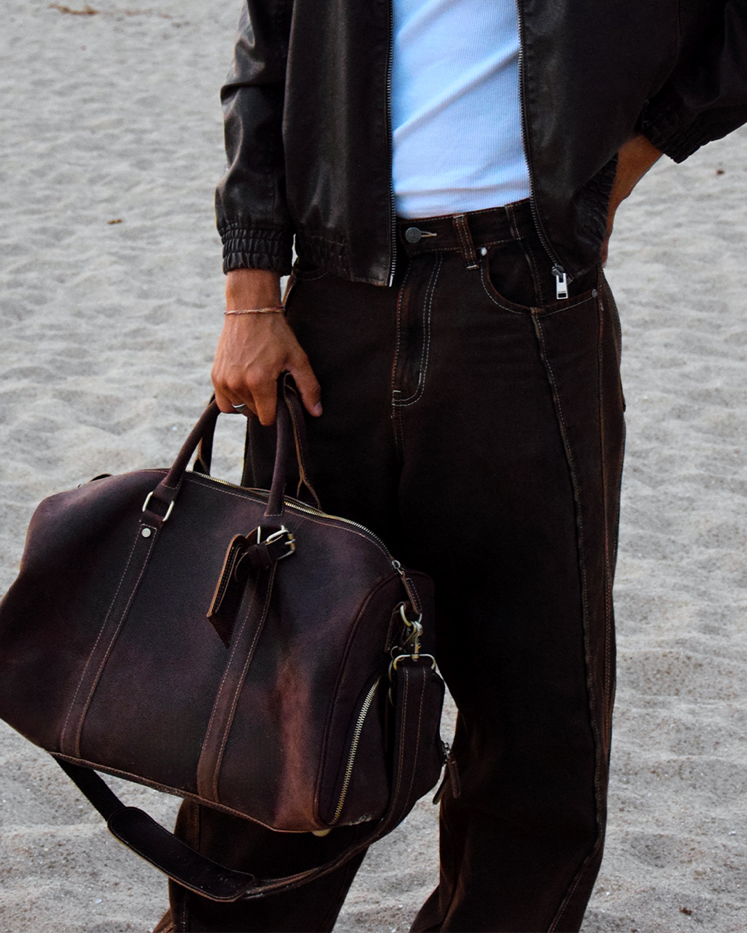 A person standing on sand holds a dark brown leather duffel bag. The person is wearing a black leather jacket, a light blue t-shirt, and dark brown jeans.