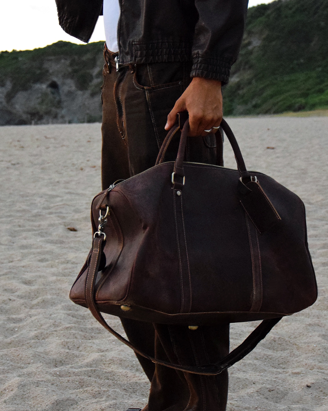 A person standing on a sandy beach holding a large dark brown leather duffel bag with top handles and a shoulder strap.