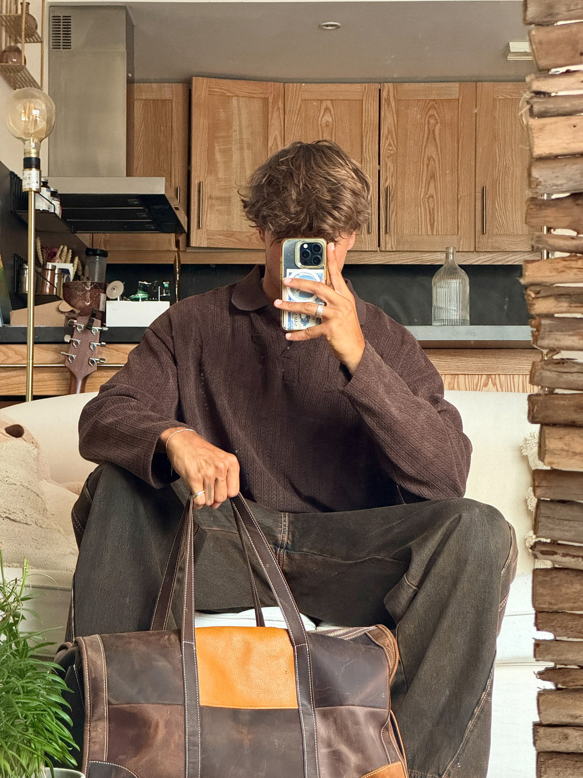 A person sitting in a home interior, holding a phone in front of their face and carrying a large brown leather duffel bag with tan and dark brown color blocking.