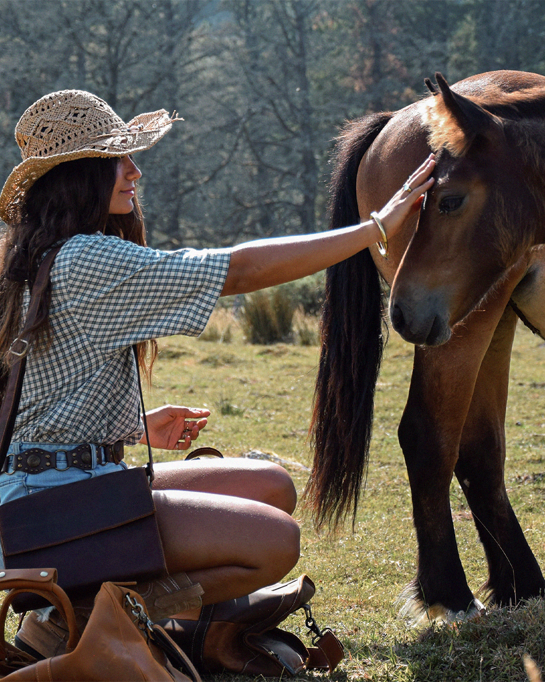 A woman in a straw cowboy hat and checkered shirt kneels beside a brown horse in an outdoor field, gently touching the horse's face. A dark messenger leather bag, duffle bag, and boots are visible.