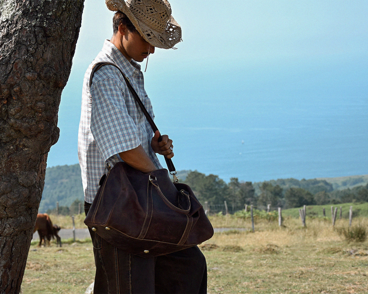 A man wearing a straw cowboy hat stands outdoors near a tree, looking down at a dark brown leather duffel bag slung over his shoulder. The background shows a scenic view of the ocean and a grassy field.