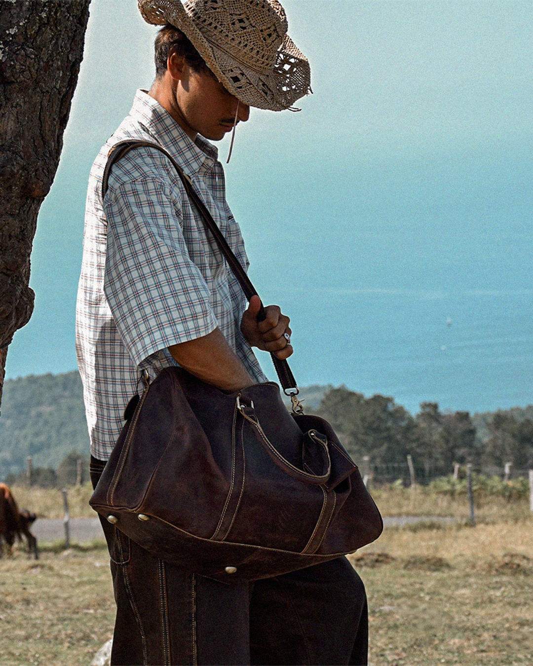 A man wearing a straw cowboy hat and a light blue-and-white plaid short-sleeve shirt carries a large dark brown leather duffel bag over his shoulder in an outdoor field with a tree trunk on the left and a blue sky in the background.