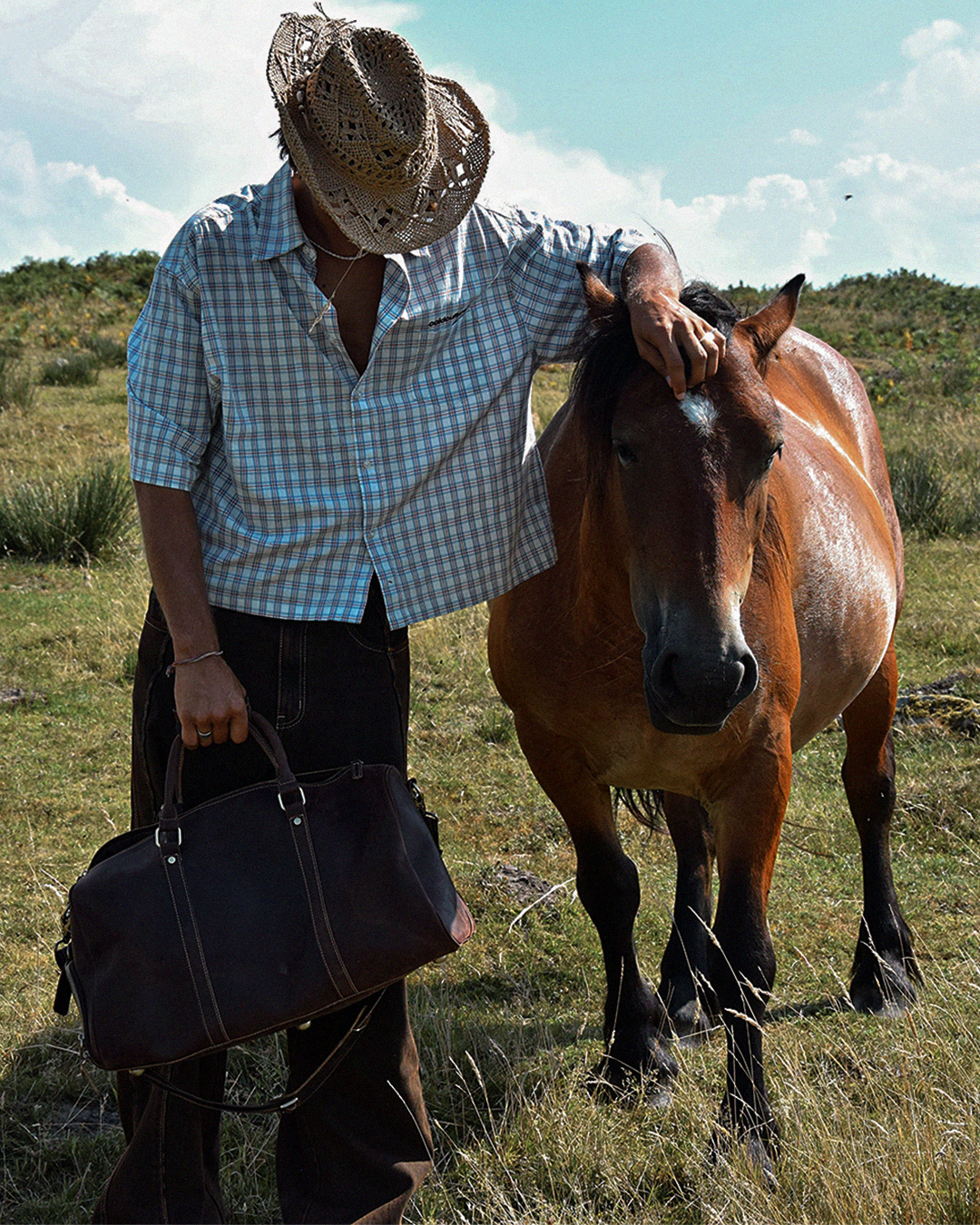 A man wearing a straw cowboy hat, a blue and white checkered shirt, and dark pants stands in a grassy field next to a brown horse, holding a dark brown leather duffel bag.