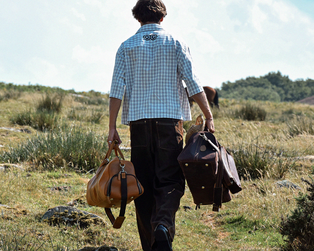 A man walking outdoors on a grassy path carrying two leather duffel bags, one tan and one dark brown, wearing a light blue and white checkered shirt.