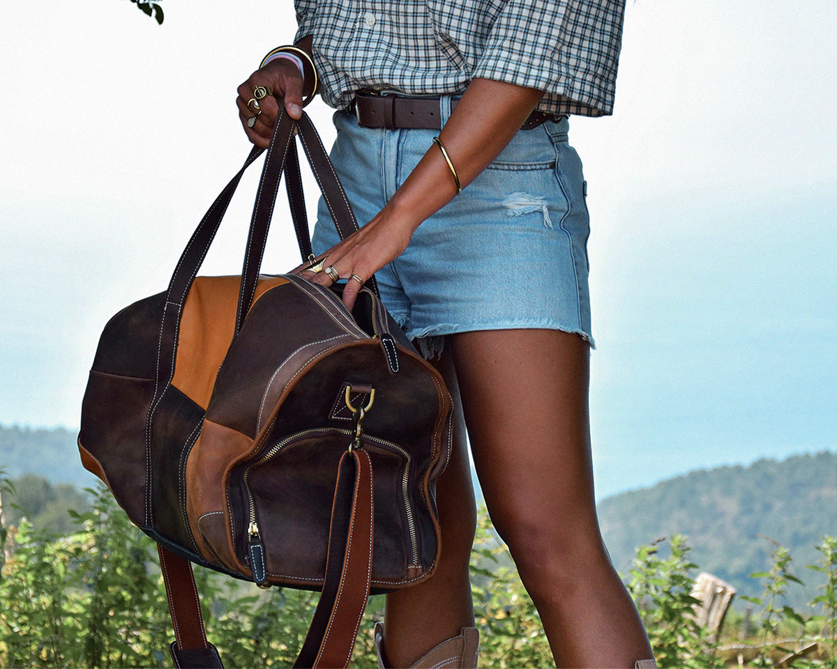 A woman outdoors carrying a large brown leather duffel bag with tan and dark brown patchwork panels, blue denim shorts, and a plaid shirt, with hills and sky in the background.