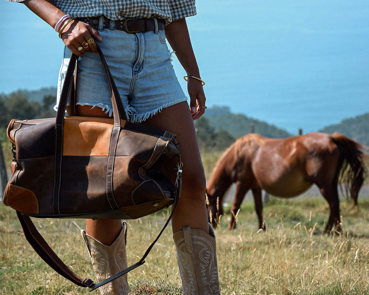 A woman outdoors carrying a large brown leather duffel bag with tan and dark brown patchwork panels. The woman is wearing denim shorts and cowboy boots, with a grazing brown horse in a grassy field under a blue sky in the background.