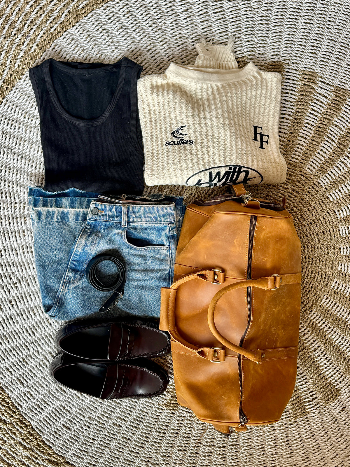 A styled flat-lay of a travel outfit on a woven circular rug, featuring a cream ribbed sweater with black logos, a black tank top, light-wash blue jeans, dark brown leather loafers, and a tan leather duffel bag.