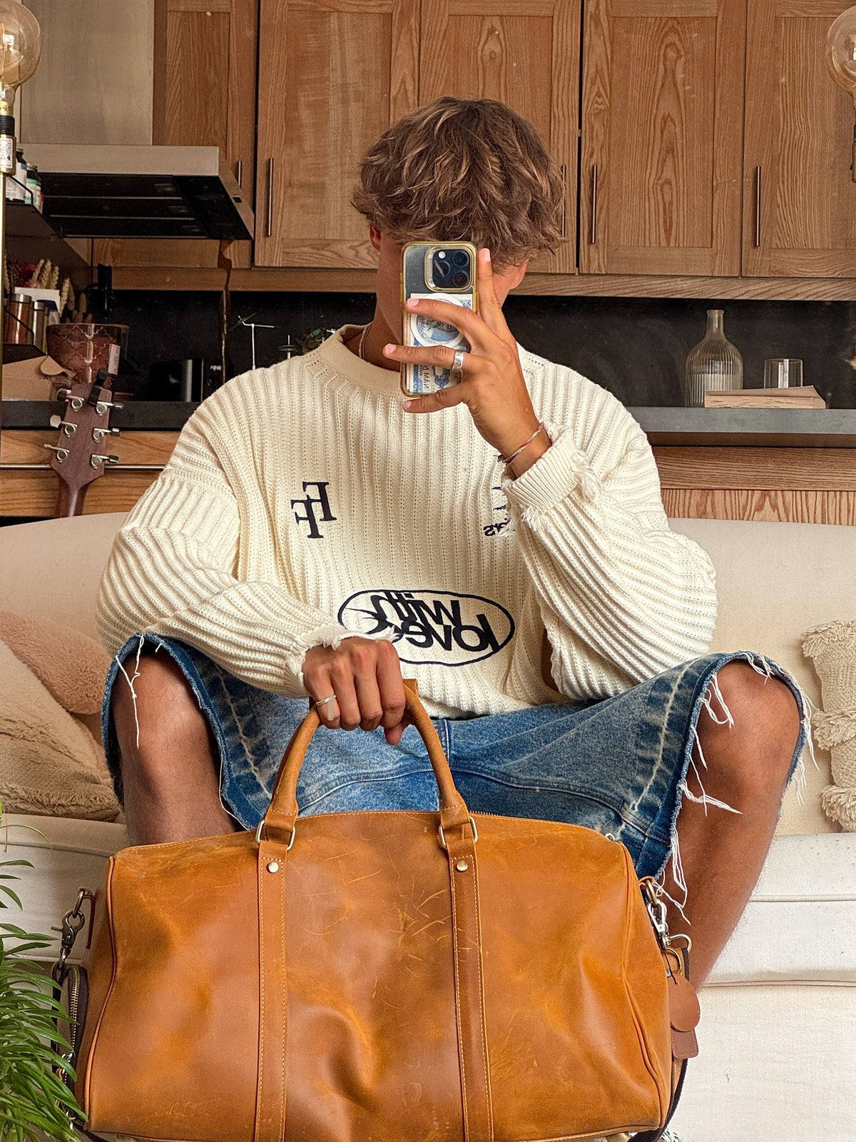 A person sitting on a couch in a kitchen setting, holding a large tan leather duffel bag in front of them. The person is wearing a cream ribbed sweater with black graphic lettering and ripped blue denim shorts.