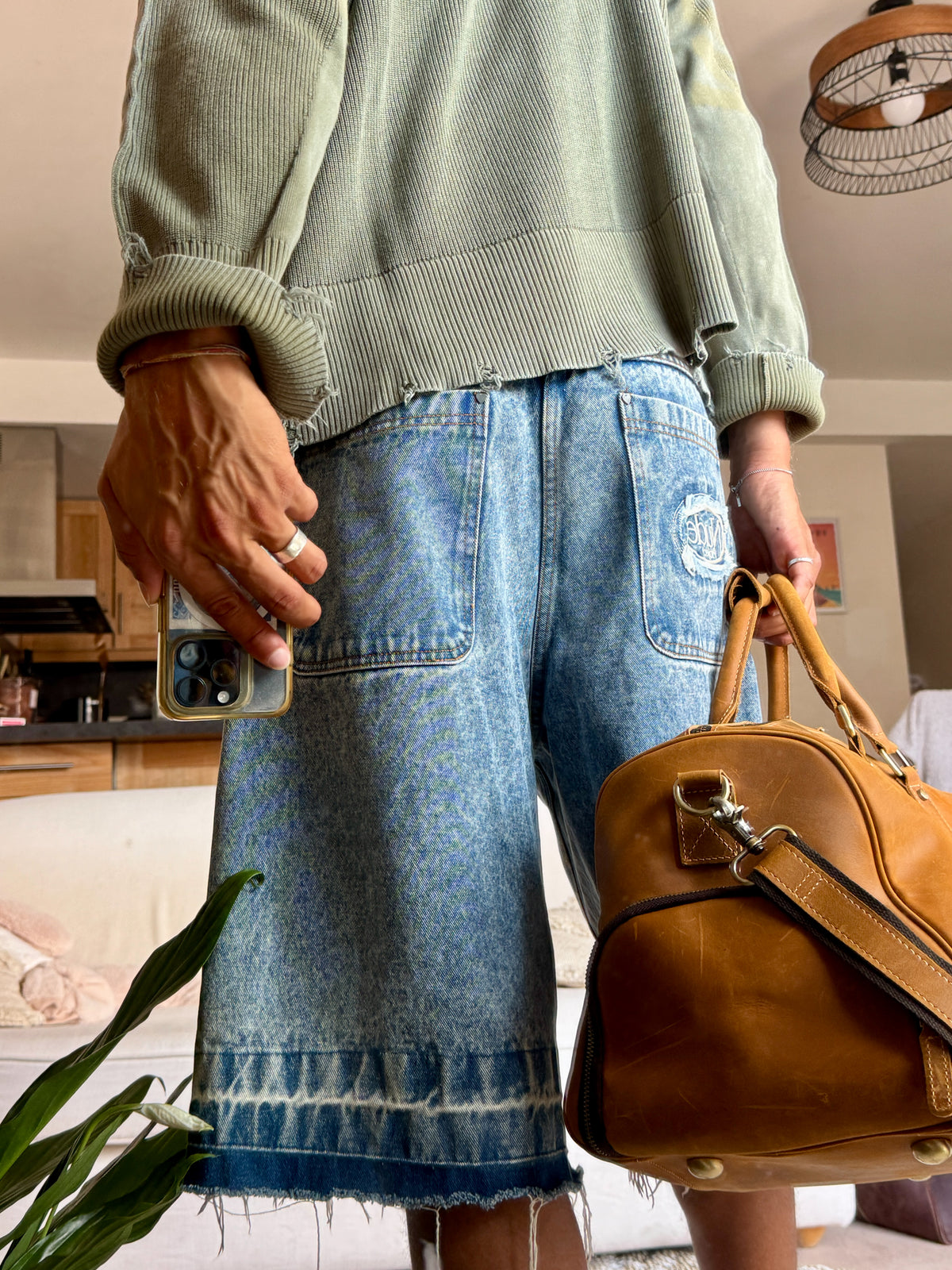 A person wearing light-wash blue denim shorts with a raw hem and a sage green ribbed sweater, holding a brown leather duffel bag in a home interior setting.