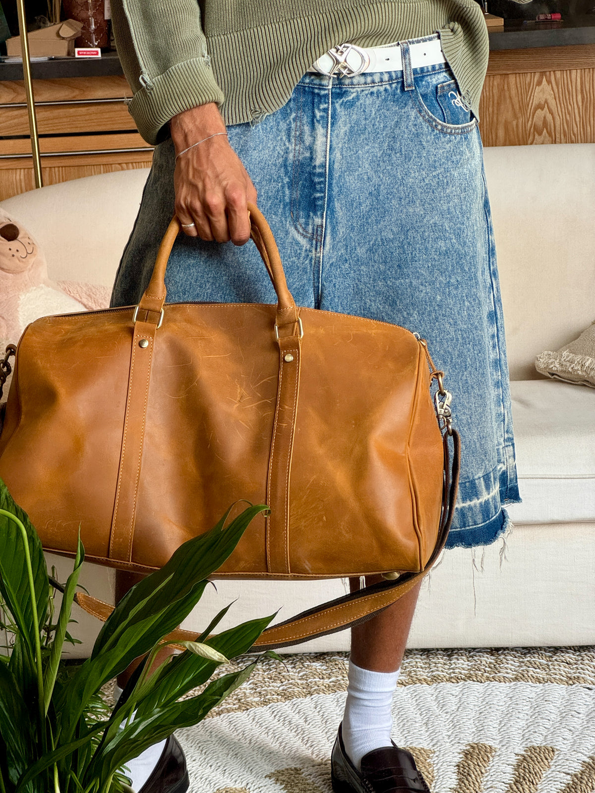 A person in a green sweater and denim skirt holds a large tan leather COFFT duffel bag with a shoulder strap in a cozy indoor setting with a sofa and plant.