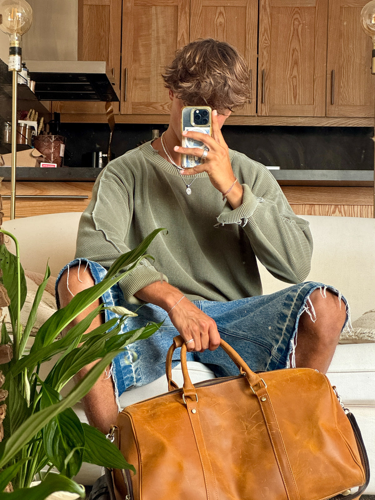 A person sitting on a white sofa in a living room setting, holding a brown leather COFFT duffel bag. The person is wearing a sage green oversized sweater and distressed blue denim shorts, holding a phone to take a mirror selfie.