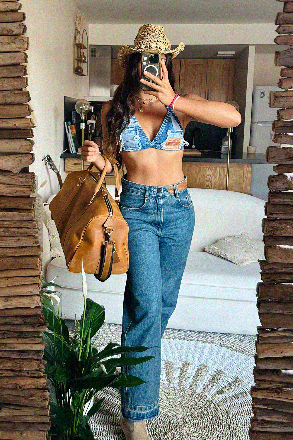 A woman takes a mirror selfie in a room, wearing a light blue denim halter top with distressed details, high-waisted blue jeans, a woven straw cowboy hat, and carrying a large tan leather duffle bag.