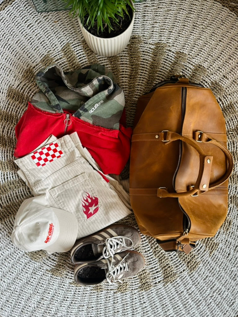 A styled flat-lay of a brown leather duffel bag alongside a red and camouflage zippered jacket, a white patterned shirt, a white cap, and gray sneakers, all arranged on a textured gray woven blanket with a small potted plant in the background.