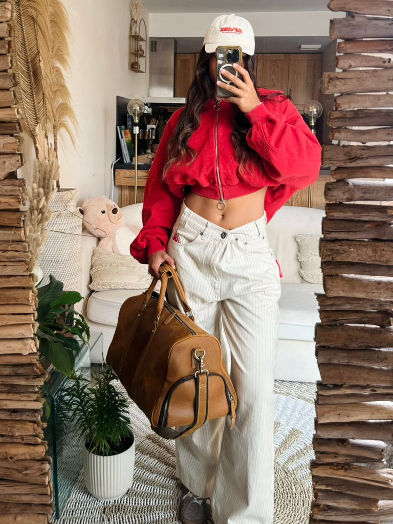 A woman takes a mirror selfie in a cozy living room setting, wearing a bright red cropped zip-up hoodie, white pinstriped trousers, a white cap, and holding a brown leather duffel bag.