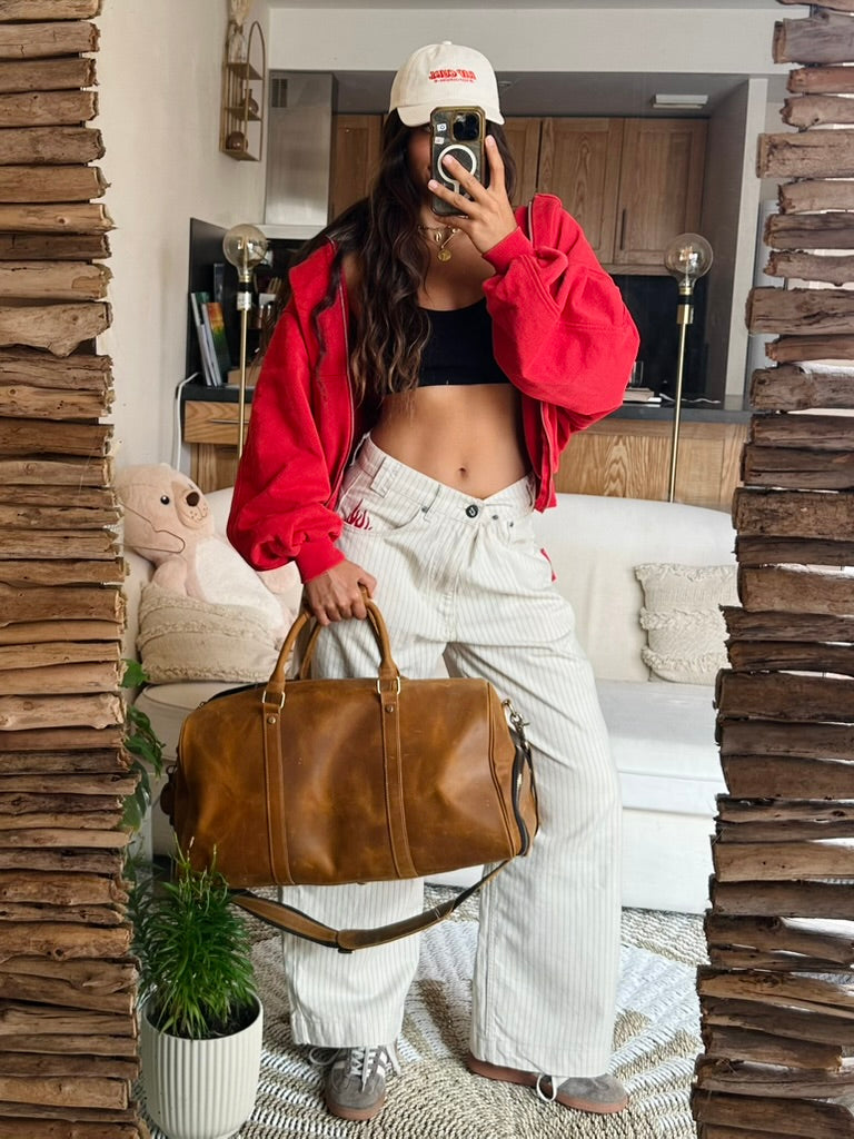 A woman takes a mirror selfie in a cozy room, wearing a bright red zip-up hoodie, white striped wide-leg pants, a black crop top, and a white cap. She is holding a brown leather duffel bag.
