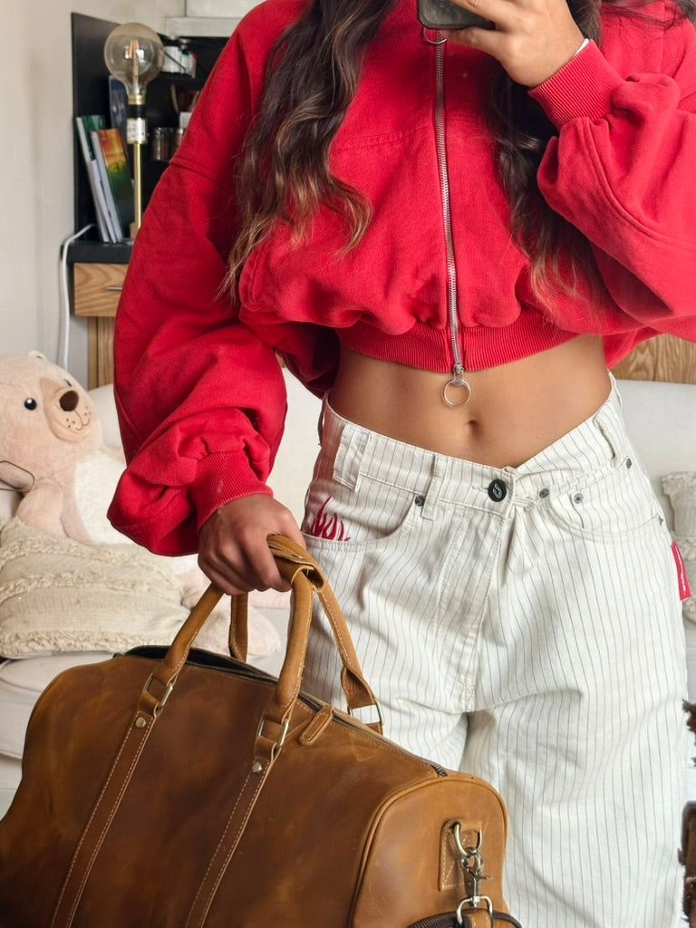 A woman takes a mirror selfie in a cozy room, wearing a bright red zip-up hoodie, white striped wide-leg pants. She is holding a brown leather duffel bag.