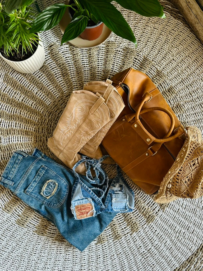 A styled flat-lay of a brown leather duffel bag, a beige western cowboy boot, blue jeans, and a straw hat arranged on a woven round rug with potted plants in the background.