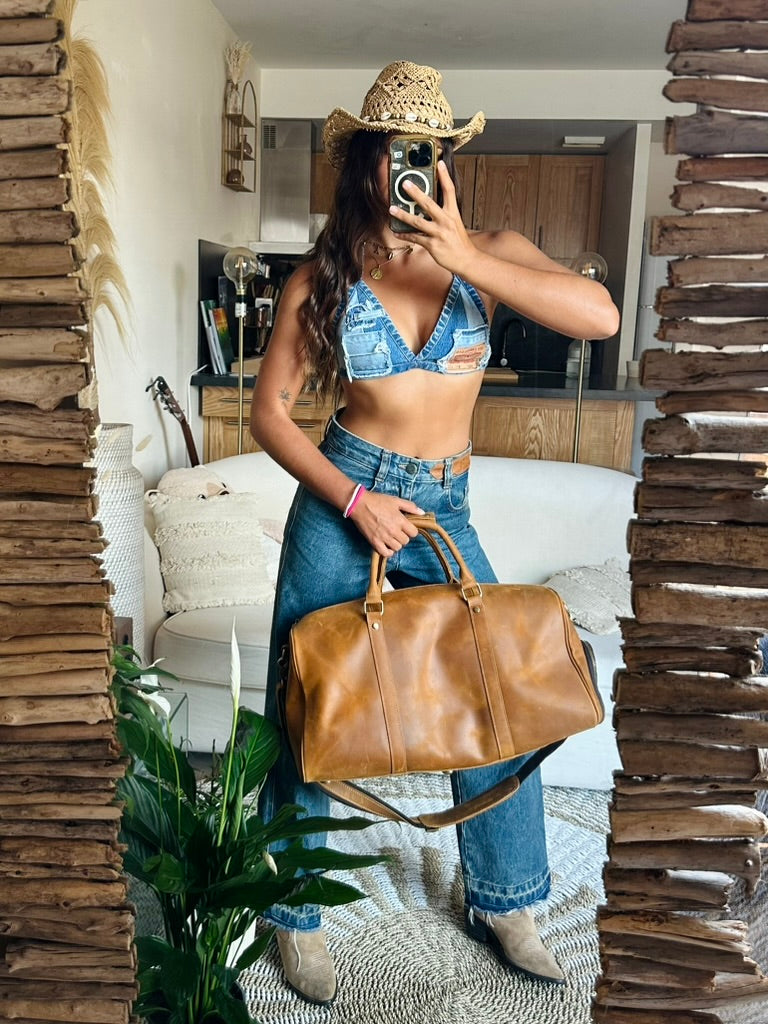 A woman takes a mirror selfie in a bedroom setting, wearing a straw cowboy hat, a denim crop top, and high-waisted blue jeans while holding a large brown leather duffel bag. 