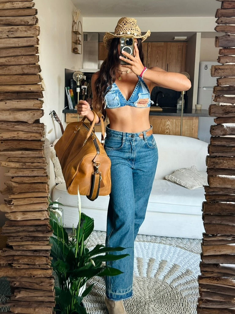 A woman takes a mirror selfie in a home setting, wearing a light blue denim triangle bralette, high-waisted blue jeans, a straw cowboy hat, and carrying a tan leather duffle bag.