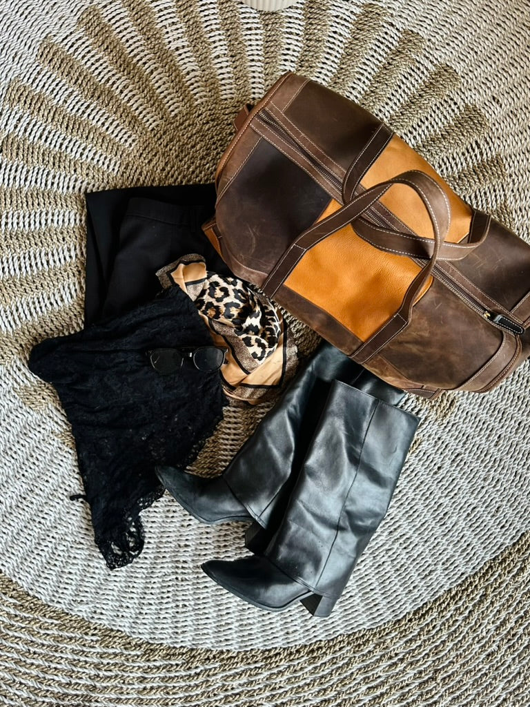 A styled flat-lay of a brown and tan patchwork leather duffel bag with black leather knee-high boots, a black knit sweater, and a leopard print scarf, arranged on a round woven straw rug.