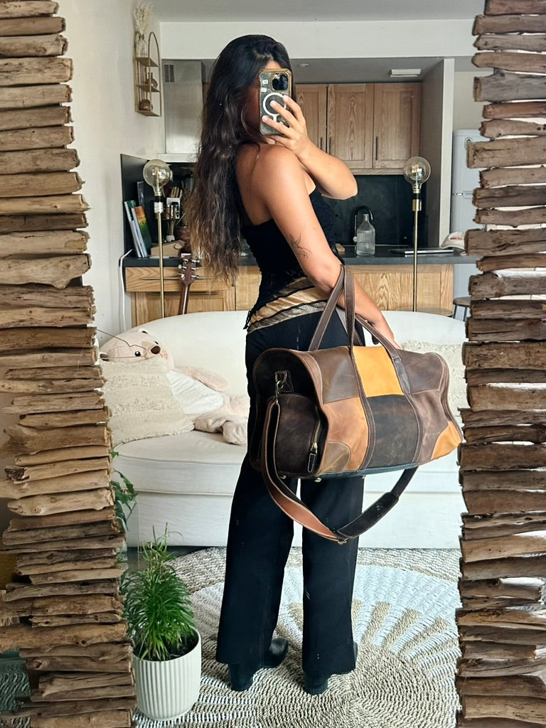 A woman takes a mirror selfie in a home setting, holding a large brown patchwork leather duffel bag with tan and mustard yellow panels. The room features rustic stacked wood pillars on either side of the mirror.
