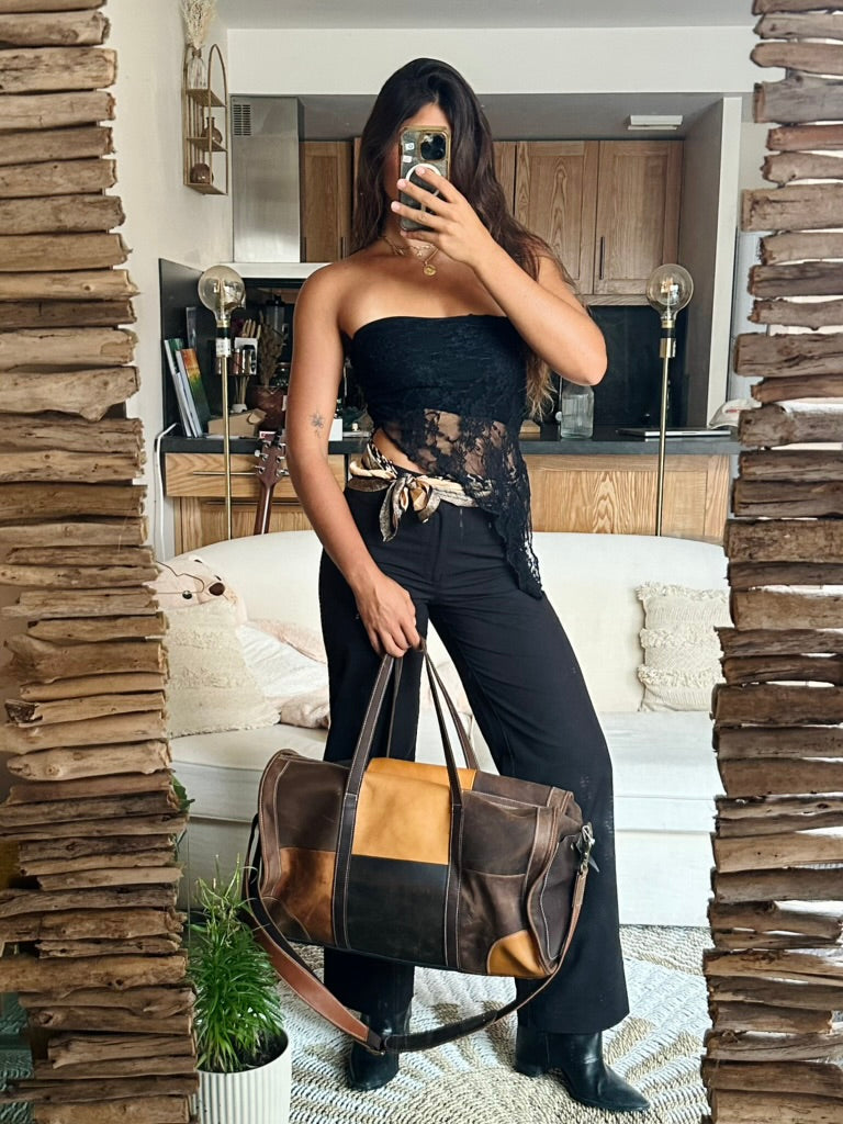 A woman takes a mirror selfie in a home setting, holding a brown and tan patchwork leather duffel bag. She is wearing a black strapless top with lace detailing and black pants.